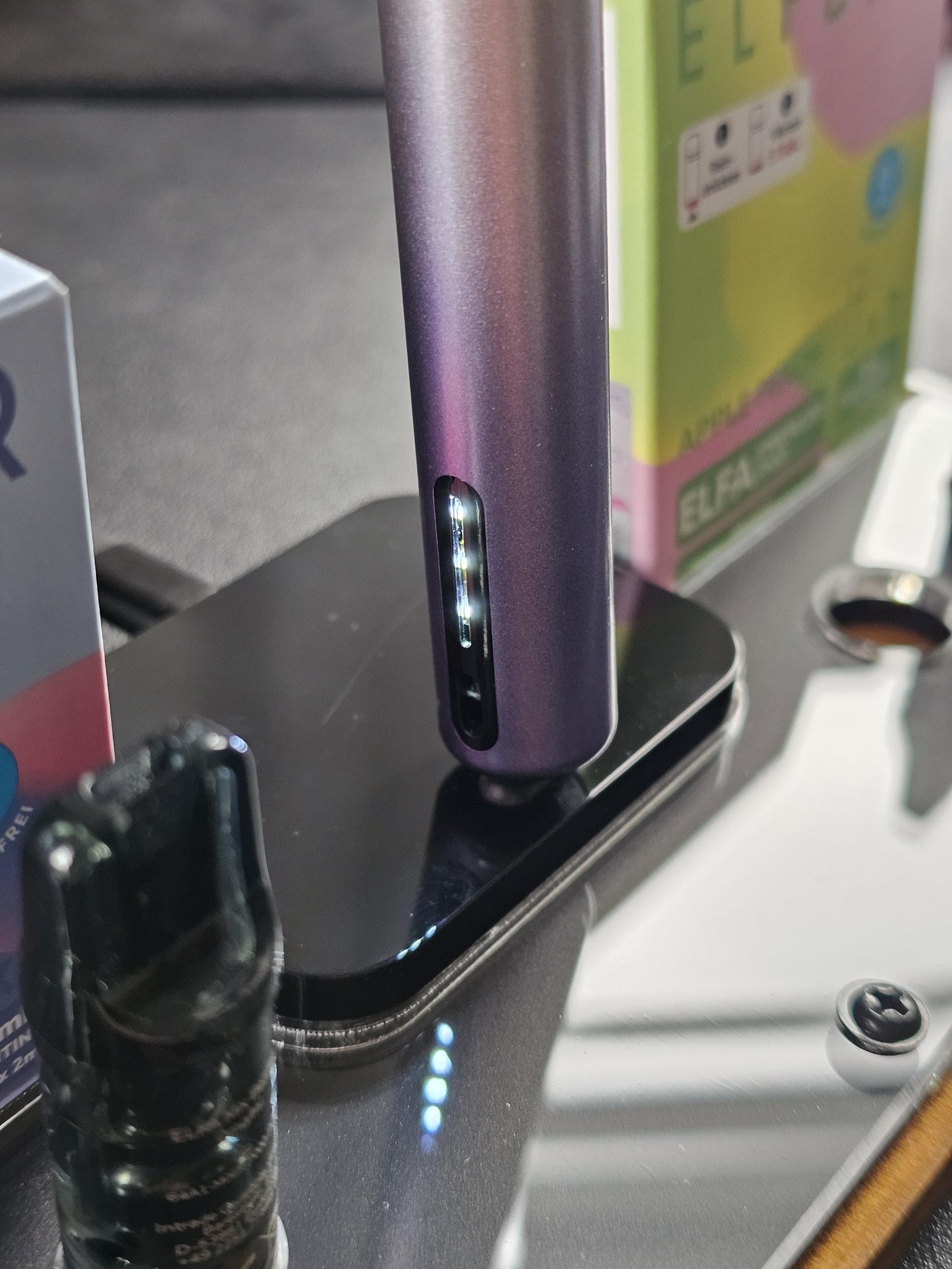 Nahaufnahme eines modernen, metallisch-farbenen Vape Pens mit Ladeschale und Verpackung im Hintergrund