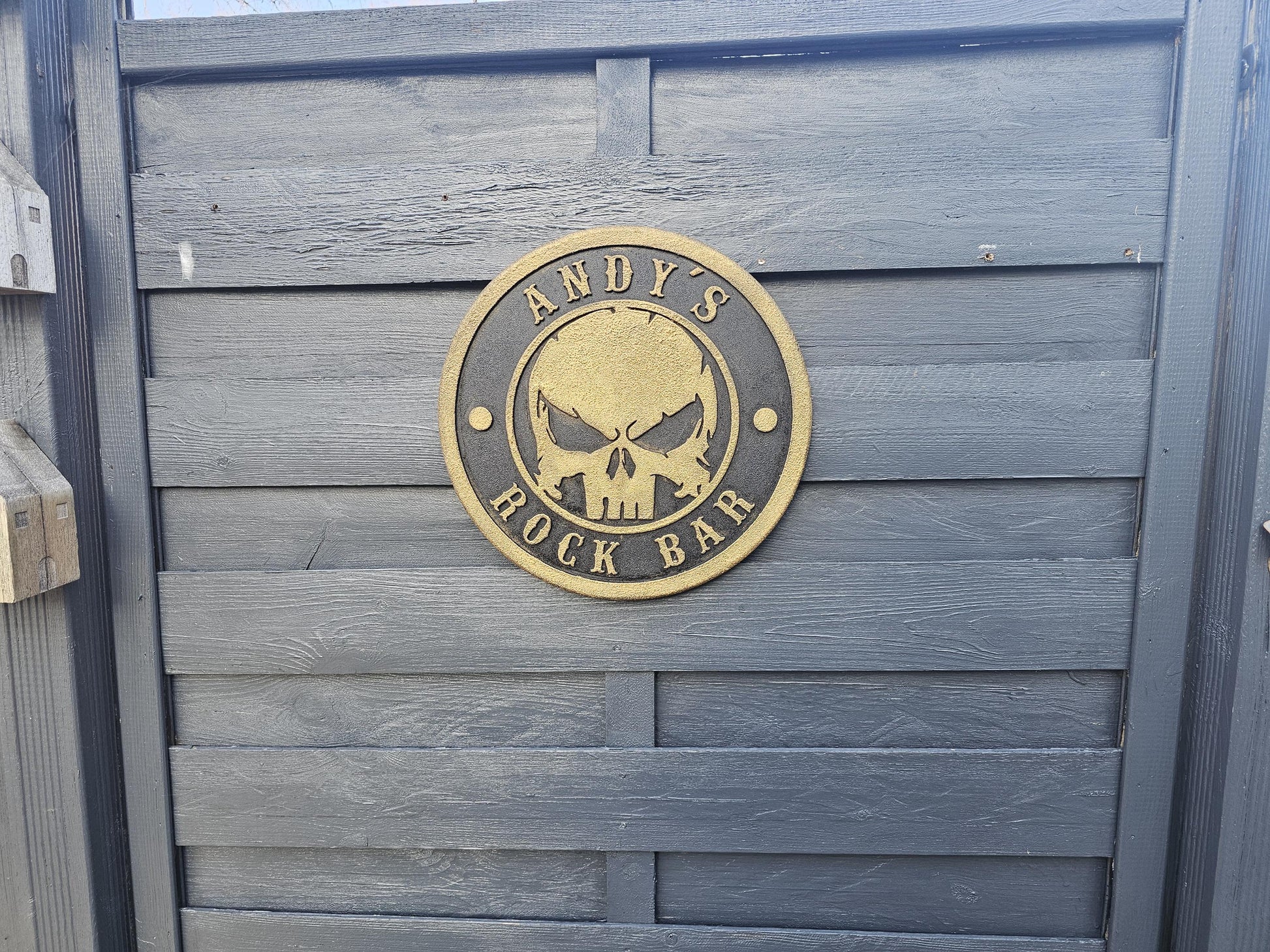 Rundes graviertes Holzschild mit Totenkopf und Text 'Andy's Rock Bar' an grauem Holzzaun.