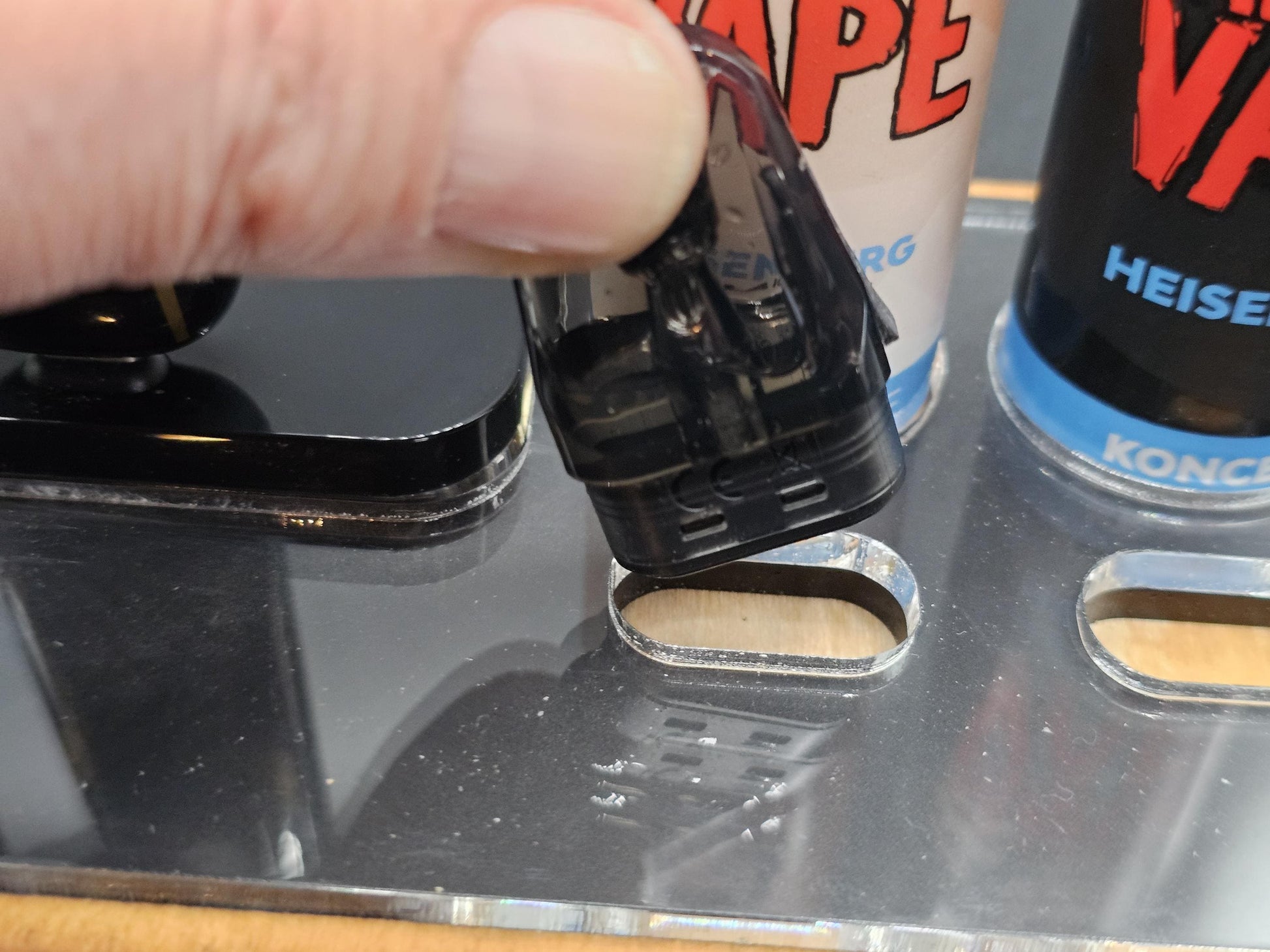 Hand hält schwarzen Pod für E-Zigarette vor Acryl-Display mit Vape-Liquids