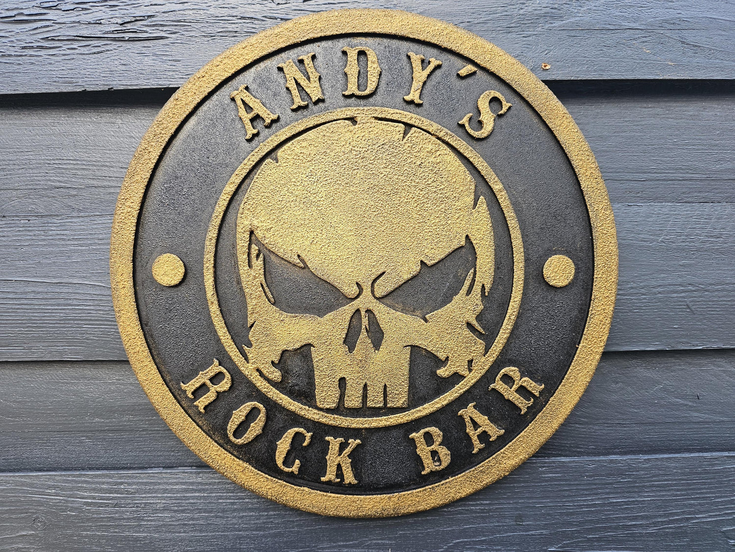 Rundes Metallschild mit goldener Totenkopfgravur und Aufschrift 'ANDY'S ROCK BAR' auf Holzwand