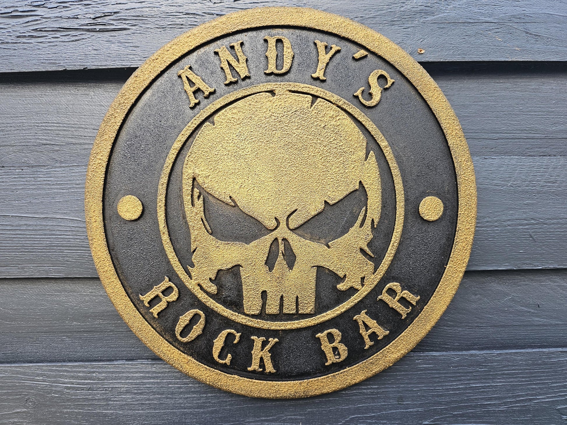 Rundes Metallschild mit goldener Totenkopfgravur und Aufschrift 'ANDY'S ROCK BAR' auf Holzwand