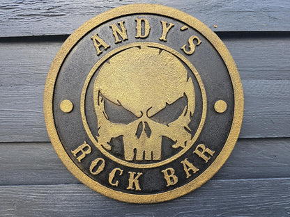 Rundes Metallschild mit goldener Totenkopfgravur und Aufschrift 'ANDY'S ROCK BAR' auf Holzwand