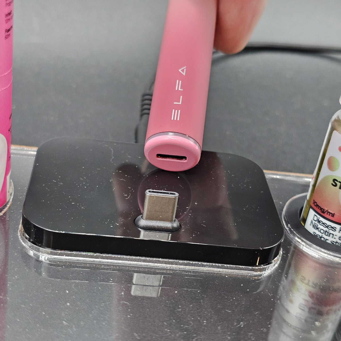 Rosa E-Zigarette mit USB-C-Anschluss auf Ladegerät, daneben Liquidflaschen